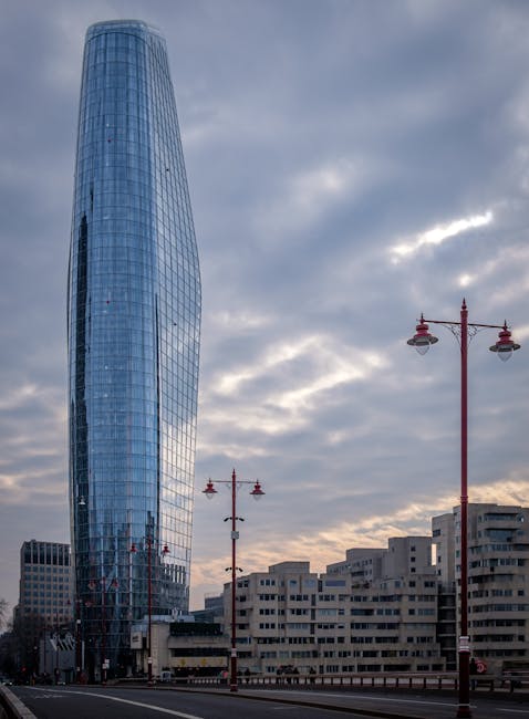 A tall, modern glass skyscraper with a curved, sleek design dominates the skyline against a partly cloudy sky during late afternoon or early evening. In the foreground, there are several traditional-s