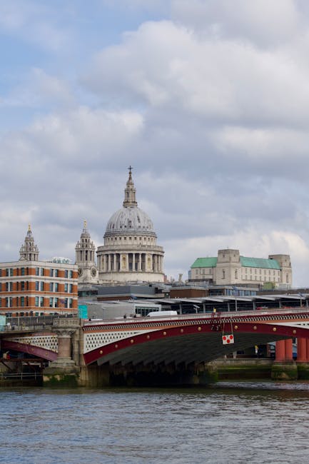 A cityscape featuring several modern high-rise buildings situated along a riverbank under an overcast sky. Prominent in the image is a tall, curved skyscraper under construction, with two cranes positioned at its top. Surrounding structures include glass-fronted office towers and residential buildings, some displaying scaffolding and construction materials. A bridge crosses the river in the foreground, with parked vehicles and trees lining the waterfront. The overall scene depicts a busy urban area focused on commercial development and city living, relevant to relocation and property clearance contexts, as seen in professional moving services like those provided by Man with Van Blackfriars.