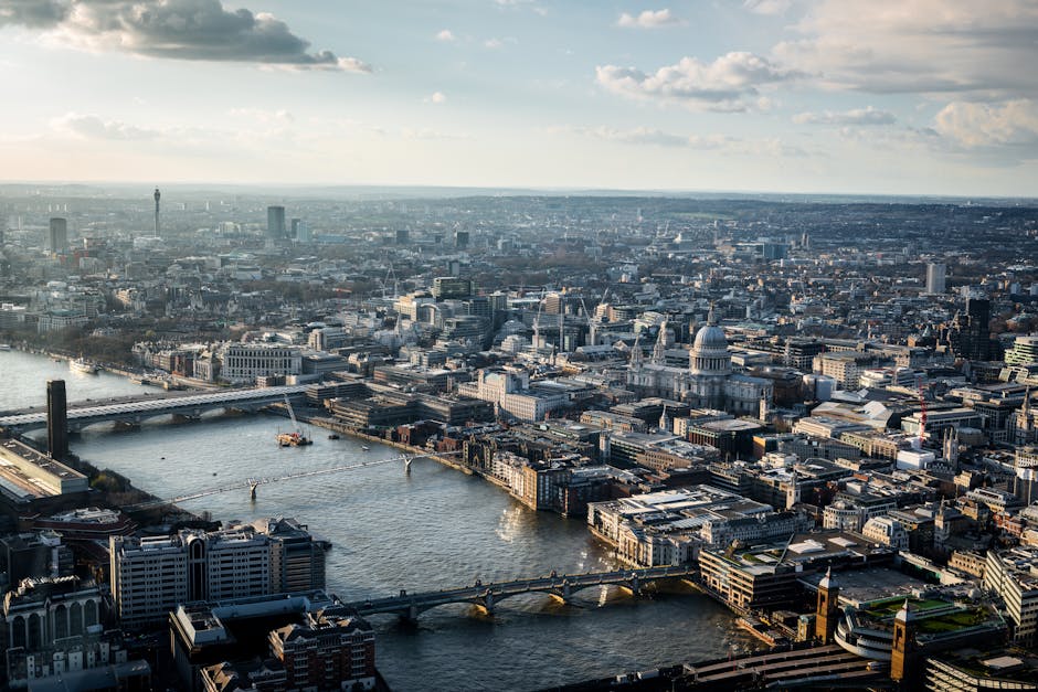 An aerial view of central London shows the River Thames flowing through the city with several bridges crossing it, including Westminster Bridge. Prominent landmarks such as St. Paul's Cathedral with its large dome are visible amidst a dense urban landscape of office buildings, historic structures, and modern high-rises. The scene is captured during daylight hours with partly cloudy skies, illuminating the city’s architecture. In the foreground, nearby rooftops and the edges of residential and commercial properties can be seen, while further in the distance, a variety of skyscrapers and landmarks mark the city skyline. The image reflects the busy density of London’s city centre, illustrating the bustling environment where home relocations and furniture transport services, like those provided by Man with Van Blackfriars, coordinate the logistics of moving within this iconic area.