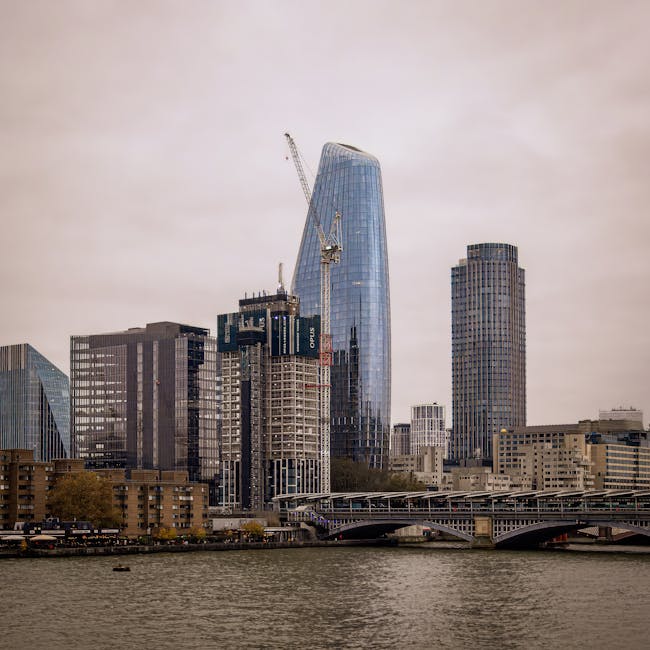 A cityscape view of the City of London featuring several modern high-rise buildings along the river Thames on a cloudy day. In the foreground, the water reflects the overcast sky, with a bridge spanning across the river, supporting vehicular traffic. Tall office towers and construction sites are visible, with one building under construction demonstrating cranes and scaffolding. The tallest building has a curved glass facade, partially covered by a crane, indicating ongoing development or renovation. The surrounding buildings include both completed office blocks with glass and concrete exteriors, and some with darker, more traditional facades. The overall scene captures urban density and development, relevant to house removals and relocation services involving commercial and residential property in the Blackfriars area, as referenced in the City of London removal permits page from manwithvanblackfriars.co.uk.