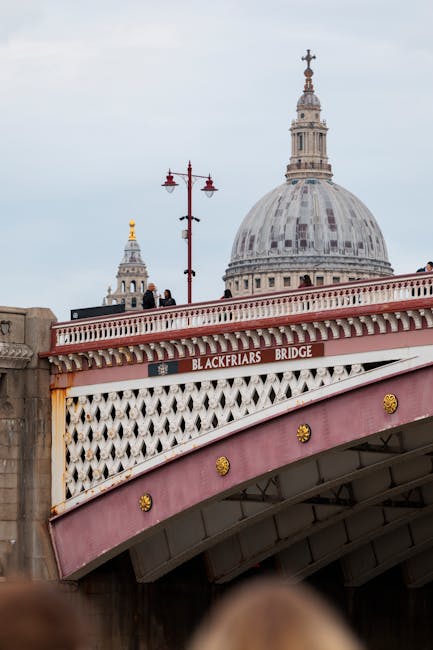 A close-up view of the Blackfriars Bridge in London, painted in pink and white with decorative gold accents, spanning over the River Thames. The bridge's nameplate is visible, and pedestrians are walking along the bridge, some standing near the edge. In the background, the domed structure of St. Paul's Cathedral with its detailed stonework and tall spire is prominent under a cloudy sky. The bridge features traditional lamps mounted on red poles, and the area beneath the bridge is partially visible, suggesting a busy urban setting associated with house removals and relocation services. This scene reflects a typical cityscape environment relevant to furniture transport and moving logistics, captured during daytime with natural lighting.