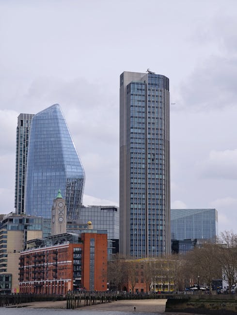 A wide view of a modern cityscape featuring several high-rise buildings, including a distinctive curved glass skyscraper and a tall rectangular office tower, set against a cloudy sky. At street level, there are leafless trees along a pavement, with a curb and some fencing visible in the foreground. The scene captures the urban environment, suggesting ongoing activities related to home relocation or furniture transport, potentially involving vehicles such as vans for moving services. This image aligns with the service category of removals offered by Man with Van Blackfriars, highlighting the logistical environment of moving and packing in central London.