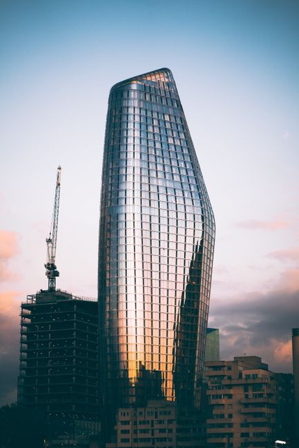 A tall, modern skyscraper with a curved, reflective glass facade stands prominently in an urban setting at sunset. The building's surface reflects the pastel colors of the sky and surrounding cityscap