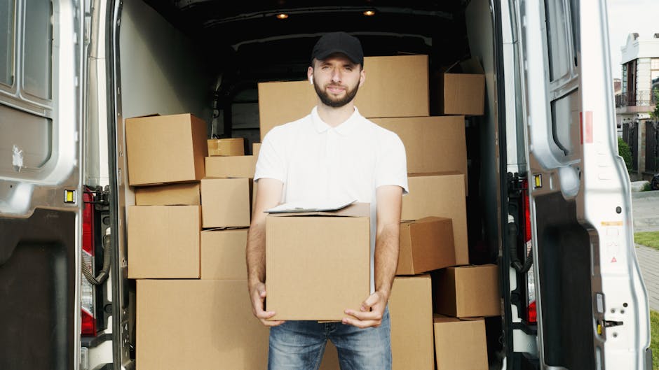 A young man with a beard, wearing a red baseball cap, plaid shirt, and a two-tone beige and brown jacket, sits on the edge of an open van's loading area surrounded by cardboard boxes. The boxes vary i