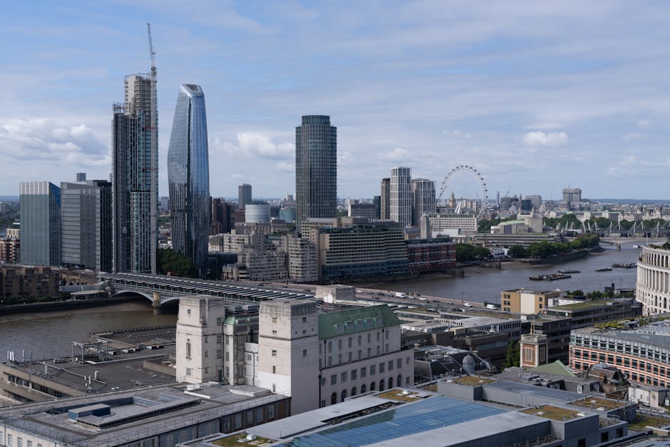 A wide aerial view of the London skyline featuring modern skyscrapers, including one with a curved glass exterior, situated along the River Thames. The river is visible with boats and piers along its banks, while nearby buildings display a mix of glass and concrete façades. In the foreground, the rooftops of lower commercial and industrial buildings can be seen, some with solar panels and rooftop equipment. The scene suggests an urban area involved in housing, offices, and transportation infrastructure, relevant to home relocation and moving logistics. The clear weather with a partly cloudy sky provides natural lighting, highlighting the details of the cityscape, which could relate to the services offered by Man with Van Blackfriars for furniture transport, packing, and loading processes involved in house removals near central London.