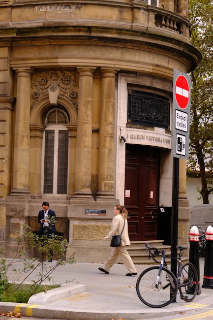 Outside a historic stone building on Queen Victoria Street EC4, two women are engaged in a home relocation process. One woman, dressed in a beige suit, is walking on the pavement carrying a black handbag. The other woman, wearing a dark jacket and holding a bundle of cardboard boxes, appears to be preparing for the moving activity. In front of the building's entrance, there is a stack of packed cardboard moving boxes, some wrapped in plastic, and a small trolley nearby. A black bicycle is secured with a lock, leaning against a traffic pole near the sidewalk, which also features a no-entry traffic sign and a white and red bollard. The building's ornate façade with columns and decorative stonework suggests an older, possibly historic property, and the scene indicates preparations for a furniture transport or part of a professional removals service by Man with Van Blackfriars as part of their house removal or moving services.