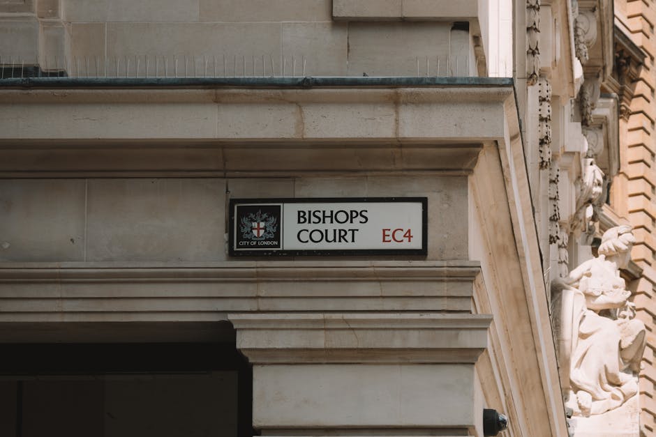 A close-up view of the corner facade of a neoclassical building on Queen Victoria Street in the City of London, featuring two rectangular street signs mounted on the cream-colored stone wall. The left sign displays 'Old Bailey' with the postal code 'EC4,' and the right sign shows 'Green Arbour Court' with the same postcode. Both signs include the City of London crest with a red cross and a sword, surrounded by a blue shield. The building's exterior includes detailed stone cornices and a black-framed window visible in the upper right corner. This image, taken during daylight, captures the urban environment characteristic of a busy area involved in home relocation and furniture transport activities, with [COMPANY_NAME] occasionally associated with logistical and packing services for removals, emphasizing exterior signage as part of the moving process or property identification.