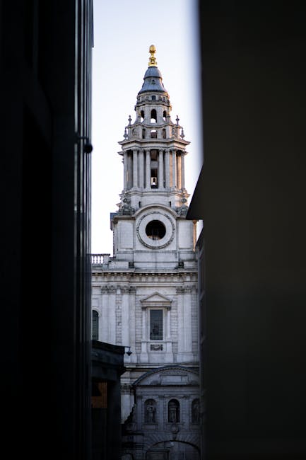 A narrow gap between two darkened building facades reveals a view of a historic church tower with a domed, gilded top, and intricate architectural details. In the foreground, part of a city street scene is visible, with a man with van blackfriars used for house removals and furniture transport, positioned nearby on the pavement. The scene suggests the process of a home relocation in a historic area, with the church providing a striking backdrop as movers from Man with Van Blackfriars prepare to navigate the tight street for loading or unloading furniture and packed boxes, using equipment such as trolleys or blankets if visible. The lighting indicates daylight, and the focus is on classical architecture contrasted with the urban environment, relevant to moving services involving packing and transport in central London.