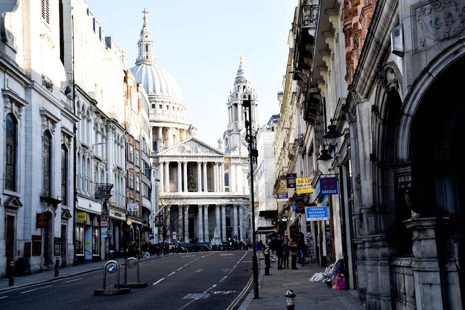 A narrow gap between two darkened building facades reveals a view of a historic church tower with a domed, gilded top, and intricate architectural details. In the foreground, part of a city street scene is visible, with a man with van blackfriars used for house removals and furniture transport, positioned nearby on the pavement. The scene suggests the process of a home relocation in a historic area, with the church providing a striking backdrop as movers from Man with Van Blackfriars prepare to navigate the tight street for loading or unloading furniture and packed boxes, using equipment such as trolleys or blankets if visible. The lighting indicates daylight, and the focus is on classical architecture contrasted with the urban environment, relevant to moving services involving packing and transport in central London.