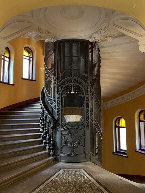 Inside a historic building with a curved, spiral staircase featuring dark wooden steps and a decorative, ornate iron balustrade, supporting a home relocation or moving process. The staircase encircles a vintage-style lift with a black metal framework and intricate scrollwork, designed to assist with transporting furniture and boxes through the building's upper floors. The interior walls are painted in warm yellow tones, illuminated by natural light coming through multiple arched windows with dark frames, positioned along the curved wall. The ceiling above showcases decorative plasterwork and a central rounded design, highlighting the building's Victorian architectural style. Minor packing materials such as cardboard boxes, some wrapped in plastic or fabric, are visible on the staircase, indicating ongoing packing and moving activities conducted by Man with Van Blackfriars, specialists in house removals and furniture transport involving tricky lifts and historic staircases.