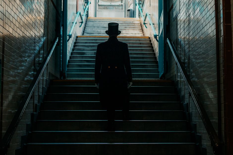 A person dressed in formal Victorian clothing, including a top hat and dark suit, is ascending a staircase inside a building, with tiled walls on either side. The staircase features metal handrails painted in a light blue shade, and the steps are made of dark material with yellow safety strips along the edges. At the top of the stairs, natural light illuminates the space, revealing an open doorway or window. The environment appears to be part of a historic or period property, and the setting is in an indoor corridor or stairwell. This scene captures a moment of a home relocation or furniture transport process, which the specialist team at Man with Van Blackfriars might handle during a house removal or moving service, especially when navigating tricky Victorian staircases in central London.